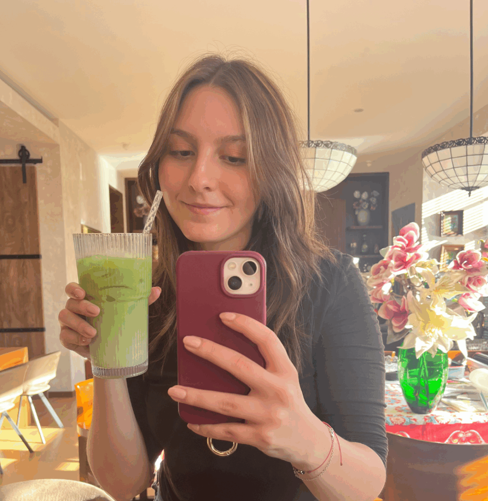 mujer sosteniendo un matcha latte en la sala de su casa, tomando una foto en un espejo.